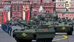 На Червоній площі рухається колона бронетехніки кольору, зокрема гусеничні бойові машини з військовослужбовцями всередині та на броні. Ліворуч видно підрозділ військових у парадній формі, які стоять шеренгою. На тлі – великі історичні будівлі з червоної цегли, прикрашені святковими банерами та емблемами.