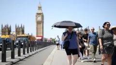 Londra'da sıcak nedeniyle şemsiye kullanan bir kadın ve etrafındakiler yürüyor. Big Ben kulesi ve Westminster arkaplanda görülüyor.
