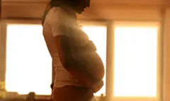 Pregnant woman standing in front of brightly backlit window Umugore utwite ahagaze imbere y'idirishya ahaturuka urumuri inyuma ye.