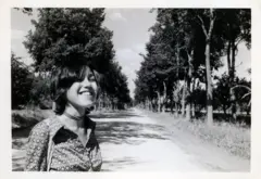 Mariona jovem em foto em preto e branco. Ela está em uma estrada cercada por árvores e sorri.