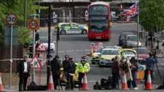مشهدٌ على الطريق يُظهر أفراداً من وسائل الإعلام يعملون بجوار طوقٍ أمني، أمام سيارات الشرطة وحافلةٍ متوقفة، بينما يرفرف العلم البريطاني في الأعلى
