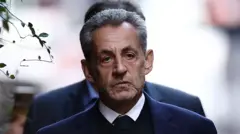 Саркози с легкой небритостью и мрачным выражением лица идет по улице.