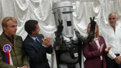 白い幕を前に壇上に並んだ候補者たち。中央の「顔がごみ箱伯爵」が黒い手袋をはめた両手でピースサインを作っている。左右の候補者たちは笑顔で拍手している
