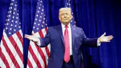 Трамп у костюмі з краваткою на тлі американських прапорів розводить руками