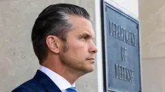 Hoton Pete Hegseth yana tsaye a kusa da wani allo mai ɗauke da rubutun "Ma'aikatar tsaro" 