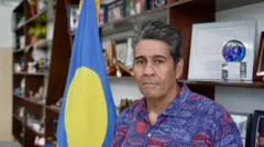 El presidente de Palaos, Surangel Whipps Jr., con una camisa azul y roja delante de la bandera del país.