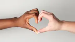 Duas mãos, uma negra e outra branca, fazem sinal de coração com os dedos sobre fundo claro