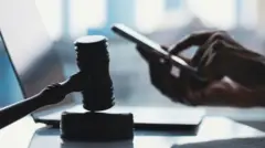Mão mexe em celular em frente a computador, com martelo da Justiça em primeiro plano