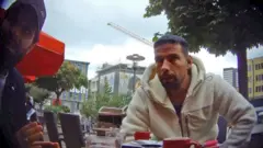 L'homme brun, barbu et aux cheveux courts, vêtu d'une veste polaire blanche, qui figurait sur les photos précédentes prises par le journaliste infiltré, discute dans un café situé sur un trottoir du centre d'Essen. Il y a des tasses de café sur la table, des gratte-ciel, des arbres et une grue en arrière-plan. Un deuxième homme - portant une casquette de baseball Gucci et une chemise bleue - avec une barbe et des cheveux foncés est visible à l'extrême gauche de la photo.