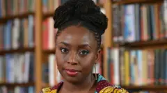 A escritora Chimamanda Ngozi Adichie está sentada em frente a uma estante de livros. Seu cabelo está preso em um coque. Ela usa brincos de ouro e uma blusa estampada em tons de vinho, turquesa e amarelo.