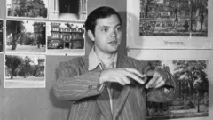 Orson Welles gesticulando em sala, onde se pode ver várias fotos de construções histórias em mural