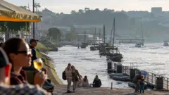 Zachód słońca w Porto na północy Portugalii. Ludzie siedzą nad brzegiem rzeki Douro. Jedna z kobiet, na pierwszym planie po lewej, trzyma kieliszek wina. Inni przechadzają się nabrzeżem i robią zdjęcia. 