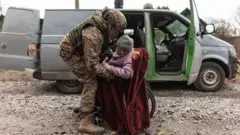 Военный поднимает пожилую женщину с инвалидной коляски во время эвакуации. На заднем плане микроавтобус с открытыми дверями 