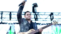 Ernesto Barajas, parado sobre un escenario, levanta su brazo derecho sosteniendo un micrófono y con una guitarra colgada.