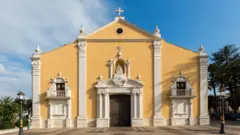 Fachada de uma igreja de estilo renascentista, com paredes amarelas e detalhes ornamentais brancos. O edifício é simétrico, com portal central encimado por uma imagem religiosa, duas pequenas varandas laterais, colunas altas e uma cruz no topo da fachada.