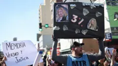 Homem sustentando um cartaz que faz referência aos supostos 3% que Karina Milei receberia pelos pagamentos da Andis à empresa Suizo Argentina