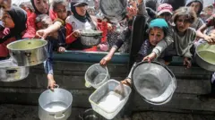 Muitas pessoas, adultos e crianças, fazendo fila para comprar comida, segurando panelas grandes e vazias.