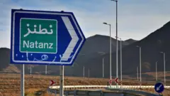 Uma placa de trânsito azul e verde que diz “Natanz” no Irã fica ao lado de uma rodovia, com uma cordilheira ao fundo
