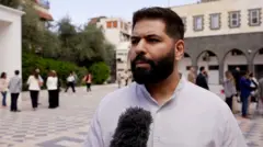 صورة تظهر شاباً بقميص مخطط بالطول يقف داخل ساحة ويتحدث للمايكروفون.