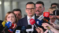 Mężczyzna z ciemnymi, krótkimi włosami ,w czarnych okularach, białej koszuli i w niebieskiej marynarce stoi w otoczeniu pięciu osób, przed nimi znajdują się mikrofony prasy
