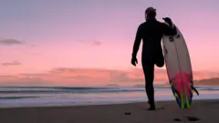 Surfista veste uma roupa de neoprene preta de corpo inteiro e luvas. Ele tem apenas uma perna e está de pé na praia, diante de um pôr do sol em tons de rosa. De costas para a câmera, olha para o mar e para a imensidão do céu. Segura uma prancha de surfe branca, rosa-choque e verde, com a ponta voltada para o céu e a outra extremidade apoiada na areia