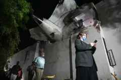 Personas caminando con mascarillas pasan por un edificio dañado que tiene en el techo restos del avión