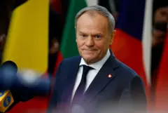 Donald Tusk em pé, rodeado por microfones da imprensa, com olhar compenetrado