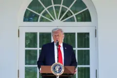 El presidente Donald Trump pronuncia un discurso durante un almuerzo en el Jardín de las Rosas de la Casa Blanca. Va vestido con traje de chaqueta azul oscuro, camisa blanca y corbata roja. Está apoyado en un atril que tiene un micrófono y un escudo de la presidencia de Estados Unidos. El fondo es una pared blanca, en medio una ventana de marcos blancos con una cortina verde.
