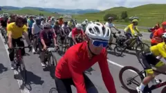 Çiçekli kırmızı üst, kask ve gözlükle bisiklet sürüyor, arkasında ve yanında çok sayıda bisikletli var. 