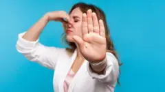 Жінка прикриває однією рукою ніс, а іншу направила на камеру