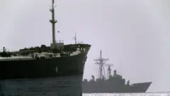 Navio de guerra americano ao fundo navega ao lado de um navio de carga no Estreito de Ormuz, durante a Operação Earnest Will