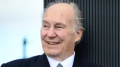 Prince Karim Aga Khan with smile wear a dark suit, white shirt and blue tie with spots
