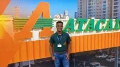 Jefferson Brito de Jesus sorrindo e usando jeans e camiseta verde, em frente ao logotipo de um supermercado