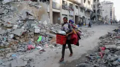 İsrail'n Gazze Şehri'nde yaşayanlara tahliye emri verilmesinin ardından, eşyalarını taşıyan bir Filistinli adam yıkıntıların arasından yürüyor elinde plastik sepet metal kap ve sırtında yorganları taşıyor. 29 Eylül 2025.