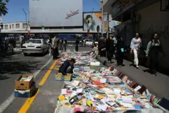 Um livreiro dispõe centenas de livros à beira de uma estrada em Teerã enquanto as pessoas passam.