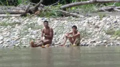 Duas pessoas estão sentadas na margem rochosa de um rio, próximas à água. Parecem nuas e estão sobre pedras lisas, entre pouca vegetação. Atrás delas, há troncos caídos e folhagem verde. O rio, em primeiro plano, tem água calma e rasa.