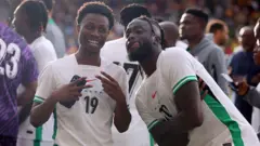 Nigerians players Tolu Arokodare bring out im tongue while posing to di camera, anoda jersey number 19 player dey beside am. Some oda palyers dey di background