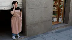 A woman stands beside a dark doorway of a closed shop in Spain
