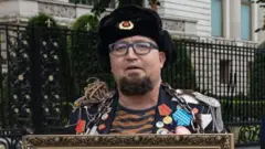 A man wearing an old-style Russian hat with a Soviet badge holds the top of a framed picture and wears a jacket crammed with medals