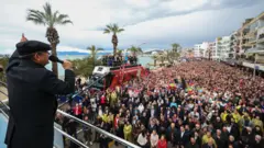 Özgür Özel kasketli bir halde otobüsün üzerinde elinde mikrofonla kalabalık bir kitleye konuşuyor