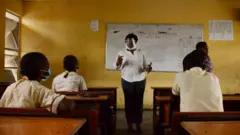 Abanyeshuri bicaye mu ishuri imbere yabo hahagaze umwarimu w'umugore