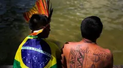 Dois indígenas de costas; um deles vestido com a bandeira do Brasil