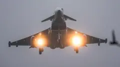 飛行する戦闘機を下から撮った写真。両翼の下についているライトが反射している