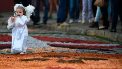 Uma criança vestida de anjo caminha sobre os tapetes de Corpus Christi.