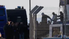 Сквозь решетку забора видно, как двое людей ведут третьего, с соединенными спереди руками (вероятно, он в наручниках). Они сходят на берег с судна, где их ждет полиция и другие люди, в гражданской одежде