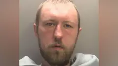 A mug shot of John Ashby who has short light brown hair and facial hair. he has a grey blanket across his front