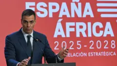Pedro Sanchez spanyol miniszterelnök beszédet mond. Mögötte egy piros, spanyol nyelvű tábla látható a következő felirattal: Spanyolország-Afrika 2025-2028.