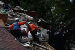 Equipos de rescate buscan entre los escombros de una estructura colapsada mientras agentes de la policía municipal permanecen cerca.