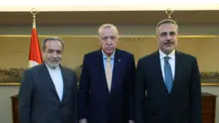 وزير الخارجية الإيراني يقف بجانب كل من الرئيس التركي رجب طيب أردوغان ووزير الخارجية التركي هاكان فيدان في مكتب رسمي وقد ظهر وراءه العلم التركي خلال زيارته لتركيا لبحث جهود نزع فتيل الحرب مع الولايات المتحدة 