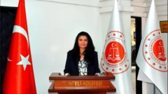 Uzun siyah saçlı, siyah takım elbiseli bir kadın kürsüden konuşma yapıyor. Sağ yanında Türkiye bayrağı var. 